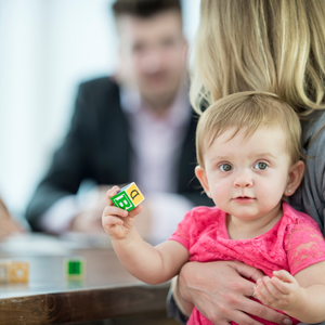 Enfant avec un cube dans les mains sur les genoux de sa mère, qui elle parle avec un adulte dans le fond.